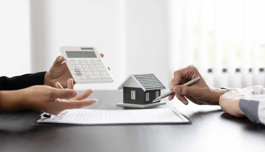 Mesa castanha onde estão duas pessoas sentadas. Na mesa estão algumas folhas, uma pequena casa de madeira e uma das pessoas segura uma calculadora. A imagem visa representar intermediários de crédito habitação