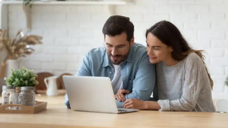 Um casal sentado numa mesa em frente ao computador. A imagem visa representar a procura de casa