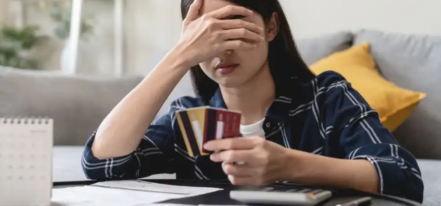 Mulher sentada no chão debruçada sobre mesa com mão na nos olhos e mão com cartões de crédito para simbolizar dívida às Finanças