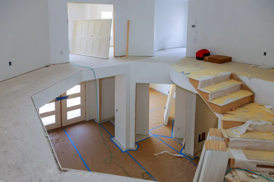 Interior de uma casa em remodelação, com escadas parcialmente desmontadas, chão protegido com papel e fita azul, e portas desmontadas encostadas às paredes.