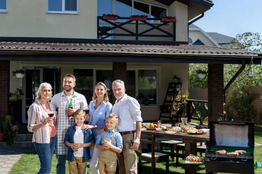 Família multigeracional reunida num churrasco no jardim de uma casa, sorrindo para a câmara, com mesa posta e grelhador ao fundo. Ambiente descontraído e ensolarado de verão.