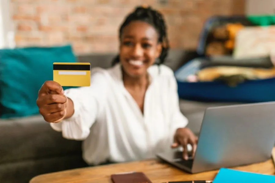 Mulher sorridente segura um cartão bancário amarelo enquanto usa um computador portátil.
