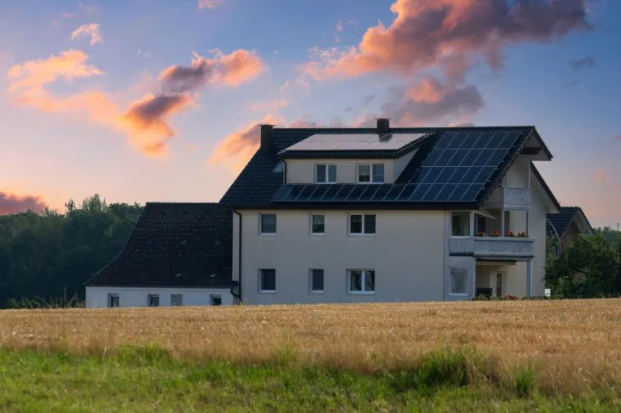 Casa moderna com painéis solares no telhado, situada numa área rural com campo de trigo dourado em primeiro plano e floresta ao fundo, ao entardecer com céu colorido em tons de rosa e azul.