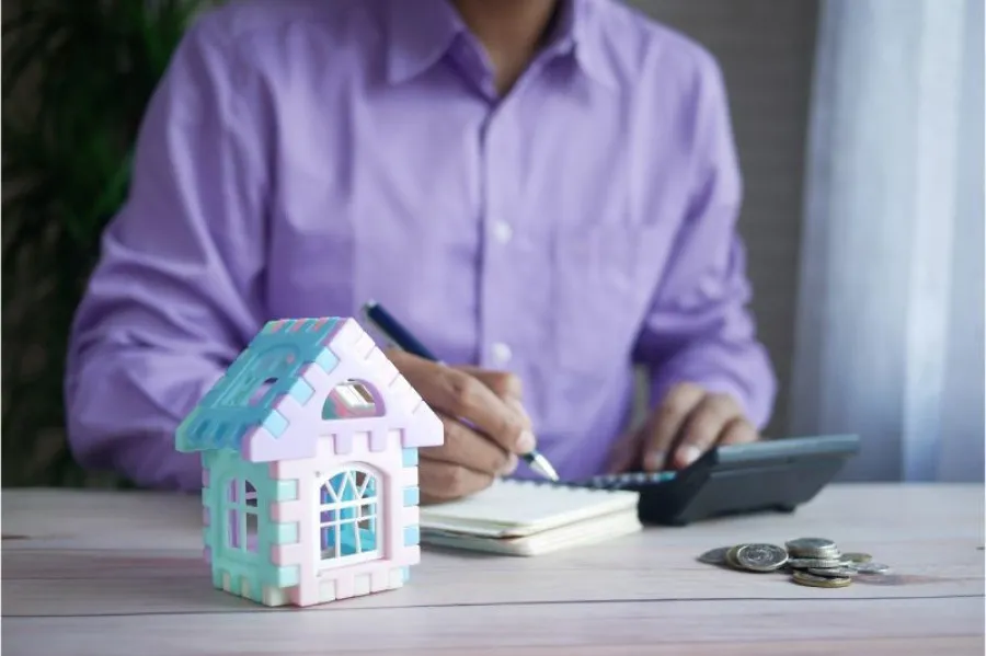 Homem de camisa roxa sentado à mesa com caneta na mão a escrever em caderno e com calculadora, maquete de casa mini e moedas na mesa