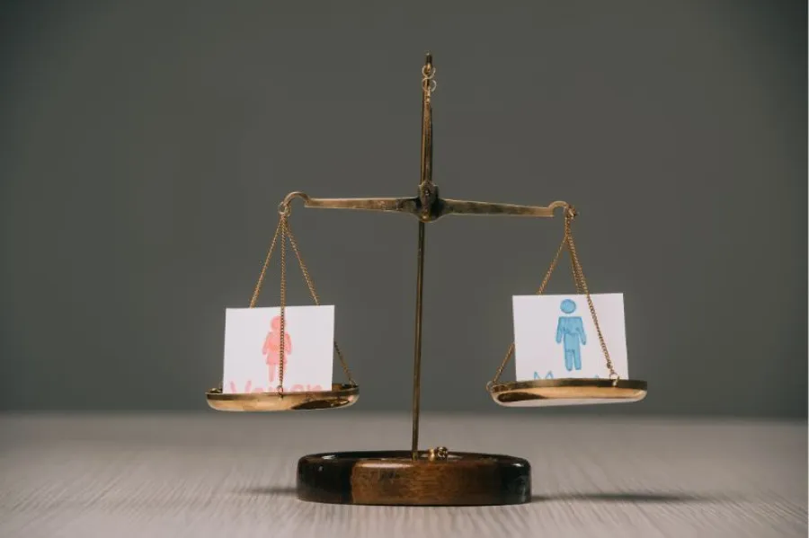 Balança de justiça com dois pratos: à esquerda, um cartão com a figura de uma mulher desenhada a vermelho; à direita, um cartão com a figura de um homem desenhada a azul. Ambos estão equilibrados, simbolizando igualdade de género.