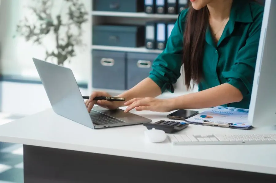Mulher a trabalhar num computador portátil com documentos, gráficos e calculadora sobre a mesa, simbolizando gestão financeira ou análise de despesas.