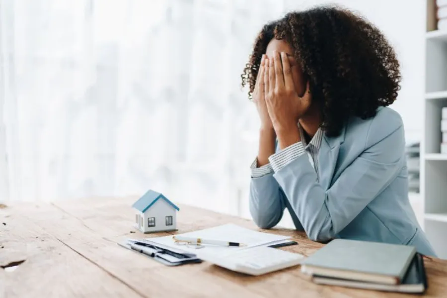 Mulher com ar preocupado a tapar o rosto com as mãos, sentada à secretária com documentos e uma miniatura de casa, representando dificuldades financeiras ou stress com crédito à habitação.