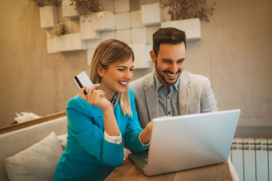 Casal jovem e sorridente usa computador portátil, com a mulher a segurar um cartão bancário, sugerindo uma compra ou operação financeira online num ambiente confortável e moderno.