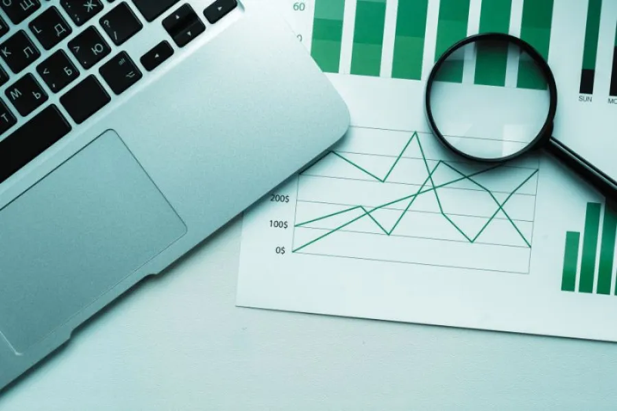 Portátil prateado parcialmente visível sobre uma mesa branca, ao lado de folhas com gráficos de barras e linhas em tons de verde. Uma lupa preta está posicionada sobre os gráficos, sugerindo análise de dados financeiros.