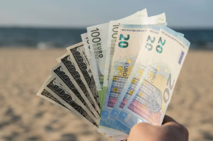 Mão a segurar várias notas de euro e dólares americanos em leque, com fundo de praia e mar desfocado ao longe, simbolizando férias e gestão de dinheiro.