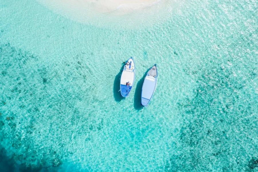 Duas embarcações pequenas flutuam lado a lado sobre águas cristalinas e azul-turquesa, próximas a uma praia de areia clara vista de cima.