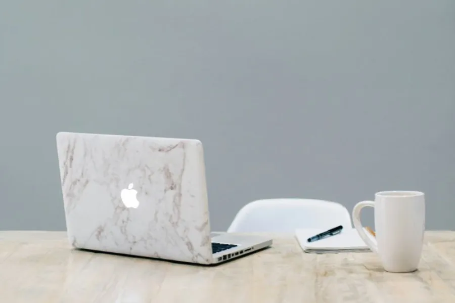 Mesa de madeira clara com um laptop com capa de mármore, um caderno com uma caneta preta em cima e uma caneca branca, em frente a uma cadeira branca, com fundo cinza liso.