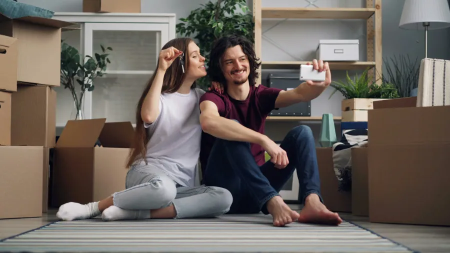 Casal jovem sentado no chão entre caixas de mudança, sorrindo enquanto tira uma selfie; a mulher segura uma chave na mão, simbolizando a compra da nova casa.