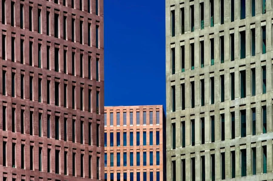 Fachadas de três edifícios modernos com janelas simétricas e cores distintas (vermelho, laranja e verde), sob um céu azul intenso.