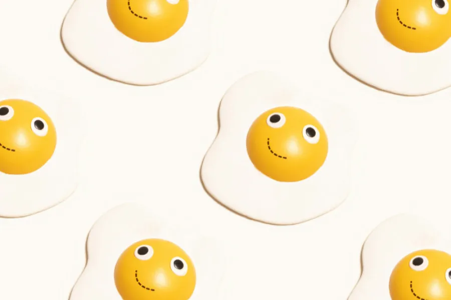 Gemas de ovo com olhos e sorriso desenhados, dispostas num fundo branco, criando um padrão divertido e alegre.