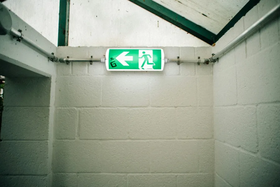 Sinal verde de saída de emergência iluminado, apontando para a esquerda, numa escadaria interior com paredes brancas de blocos de cimento.