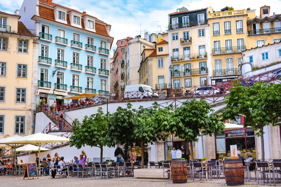 Vista de uma esplanada com árvores e barris de vinho na Praça do Município, em Lisboa, rodeada por prédios típicos e coloridos com escadaria ao fundo.