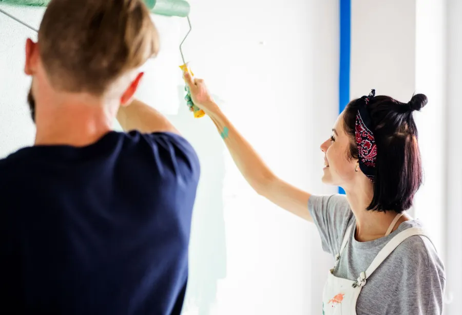 Casal jovem a pintar uma parede de branco e verde claro, juntos, durante obras de renovação numa casa.