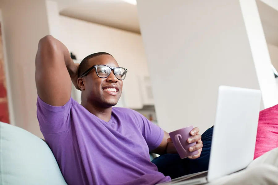 Homem sorridente com óculos, camisa roxa e caneca roxa na mão, sentado no sofá com portátil no colo em ambiente caseiro luminoso