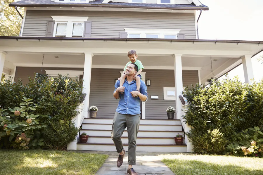 Homem sorridente com camisa azul e calças bege com um menino de camisa verde aos ombros, à frente de uma casa cinzenta com jardim e vasos de flores