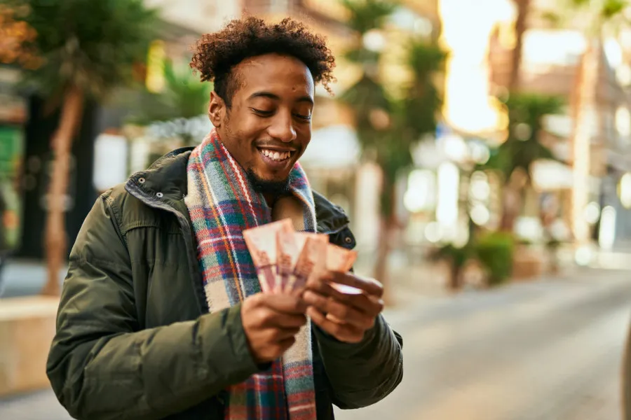 Jovem com cabelo cacheado, casaco verde e cachecol xadrez multicolorido, sorri feliz na rua enquanto segura notas de dinheiro