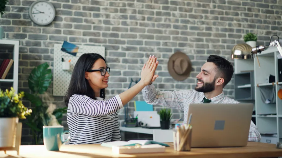 Duas pessoas numa sala de trabalho trocam um high-five enquanto uma delas está sentada ao portátil, com mesa, café e decoração de escritório ao fundo.