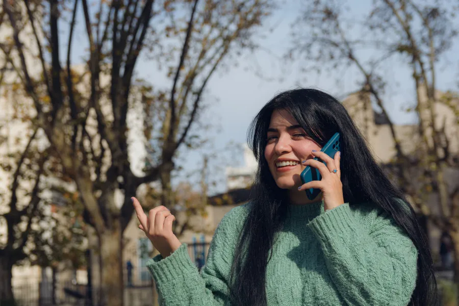 Mulher com cabelo comprido e camisola verde fala ao telemóvel ao ar livre, com árvores desfocadas ao fundo.