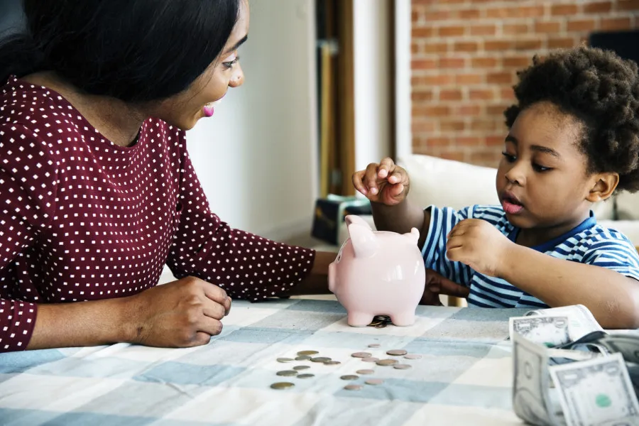 Mãe sorridente e filho pequeno brincam com mealheiro e moedas sobre toalha quadriculada.