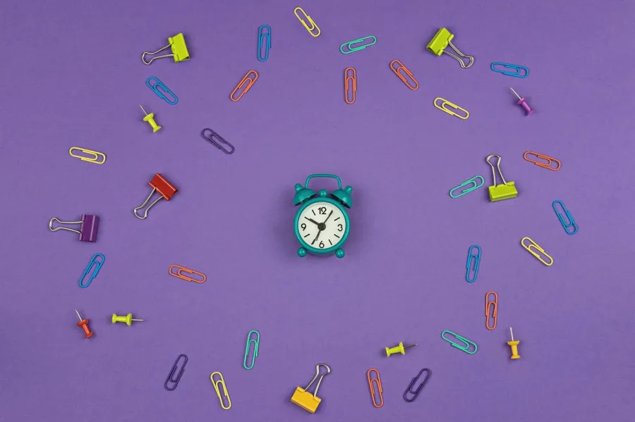 Relógio verde ao centro, com artigos de papelaria à volta, num fundo roxo.