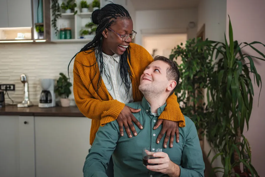 Mulher com casaco amarelo abraça homem sentada à mesa de uma cozinha, com plantas e bancada ao fundo.