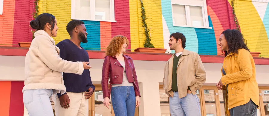 Grupo diverso de cinco pessoas conversa e sorri em frente a uma parede colorida.