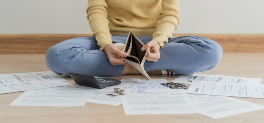 Mulher sentada em chão com folhas espalhadas, calculadora, moedas, cartões e a abrir carteira vazia