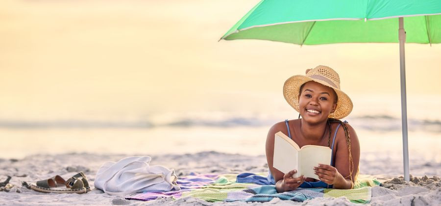 Senhora deitada em toalha na praia a ler livro sob chapéu de sol simbolizando férias poupadas em destinos baratos